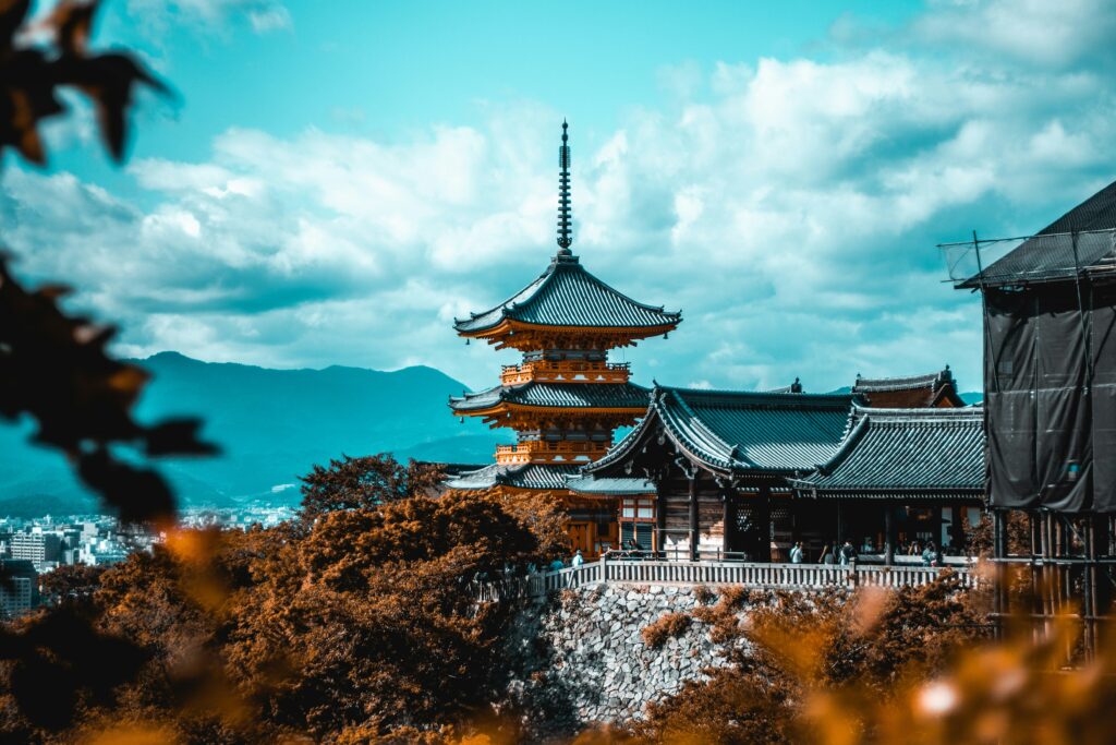 Kiyomizu-dera's three-storey vermilion pagoda in Kyoto, framed by autumn leaves with mountains behind