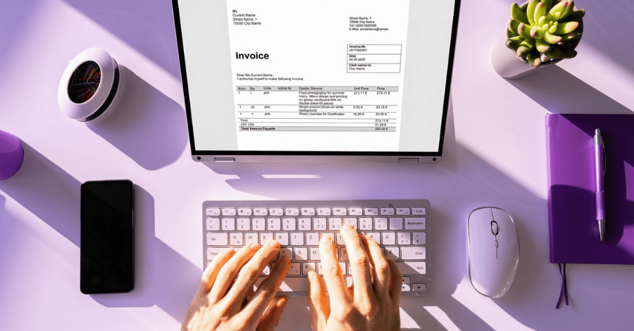 Hands typing on a laptop showing an invoice, on a purple desk with a notebook, phone and mouse