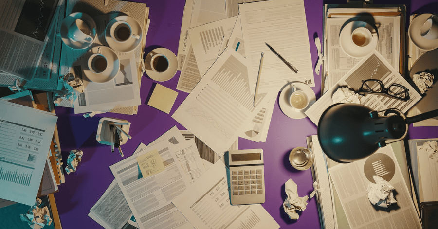 Overhead view of a purple desk covered in business paperwork, charts, a calculator, coffee cups and reading glasses