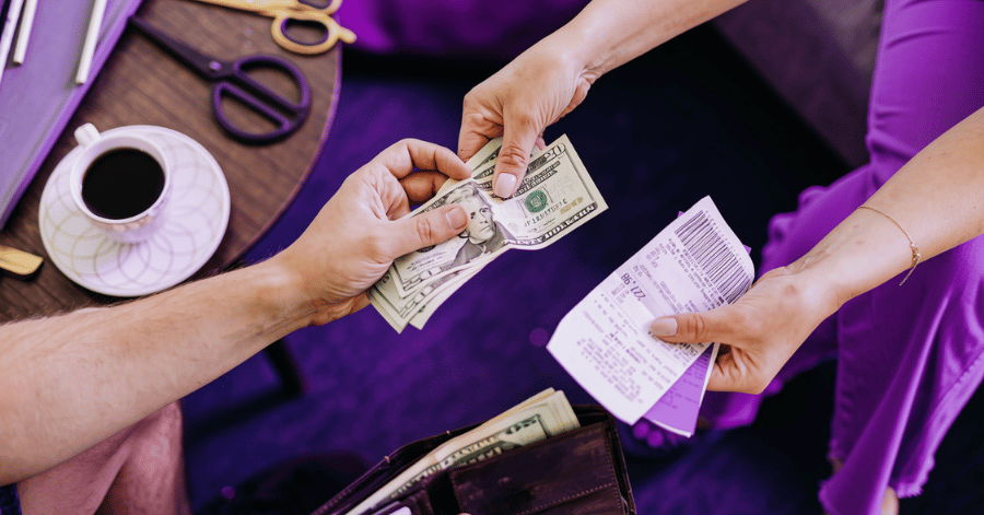 Two people exchanging banknotes and paper receipts across a desk beside a cup of coffee