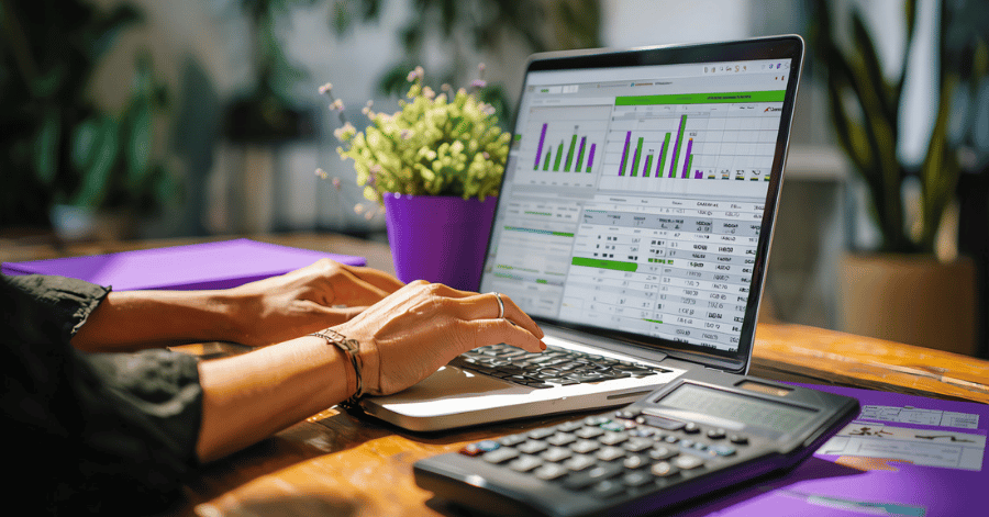 Hands typing on a laptop showing an accounting spreadsheet with bar charts, beside a desk calculator