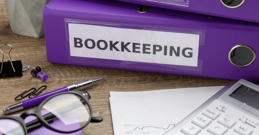 Purple ring binder labelled Bookkeeping on a wooden desk with a calculator, glasses and printed reports