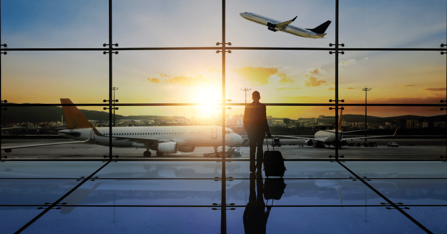 Business traveller with a wheeled suitcase watching planes from an airport terminal window at sunset