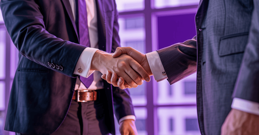 Two people in business suits shaking hands in front of an office window, lit in purple