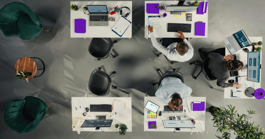 Overhead view of an open-plan office with staff working at white desks with monitors, laptops and purple folders