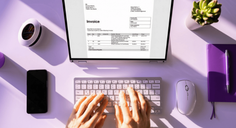 Hands typing on a laptop showing an invoice, on a purple desk with a notebook, phone and mouse