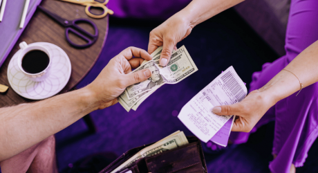 Two people exchanging banknotes and paper receipts across a desk beside a cup of coffee