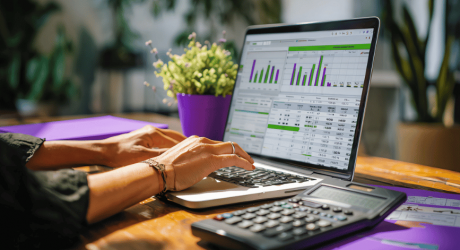 Hands typing on a laptop showing an accounting spreadsheet with bar charts, beside a desk calculator