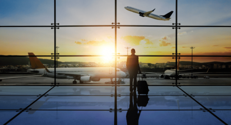 Business traveller with a wheeled suitcase watching planes from an airport terminal window at sunset