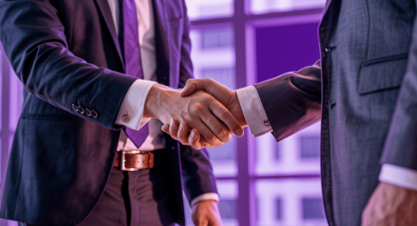 Two people in business suits shaking hands in front of an office window, lit in purple