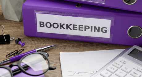 Purple ring binder labelled Bookkeeping on a wooden desk with a calculator, glasses and printed reports