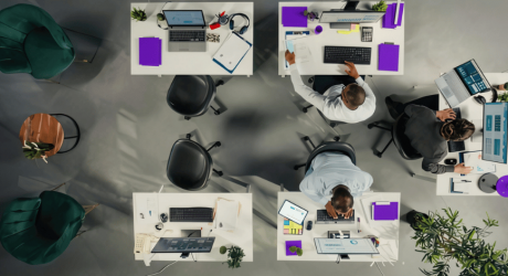 Overhead view of an open-plan office with staff working at white desks with monitors, laptops and purple folders