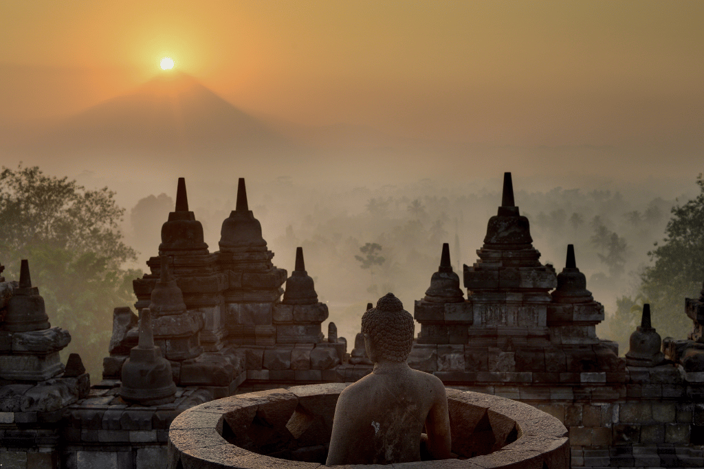 A stone Buddha and Borobudur's stupas at sunrise, with Mount Merapi silhouetted behind