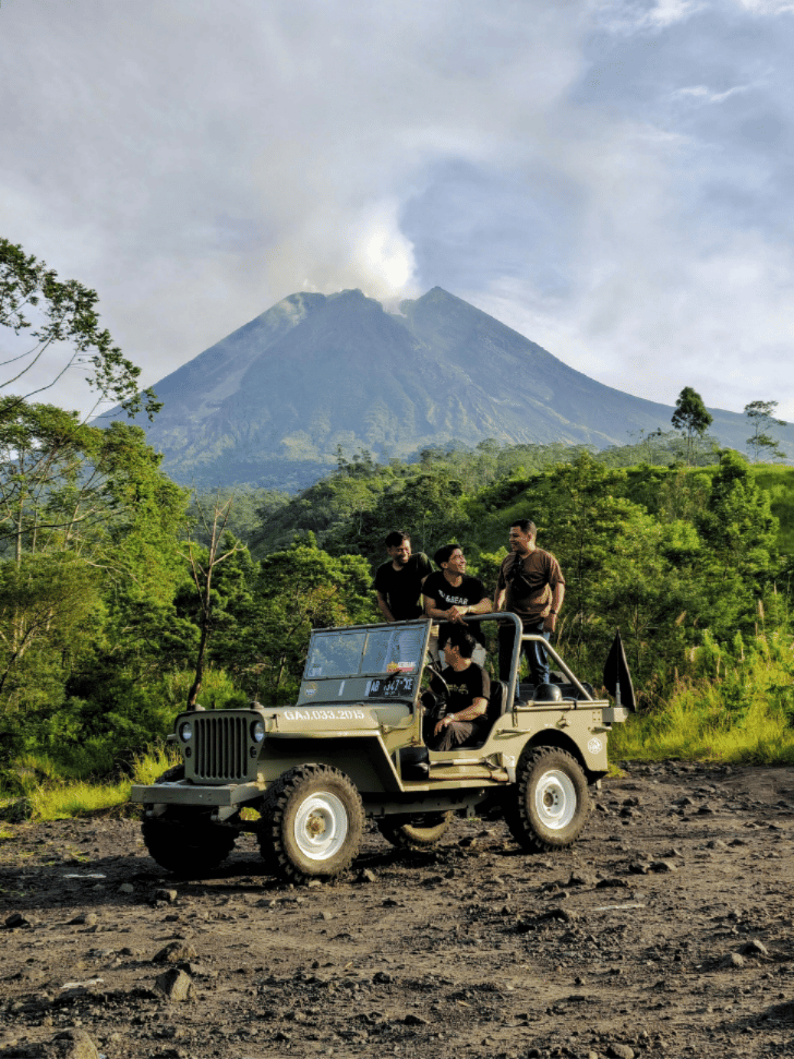 A vintage jeep and tourists on a rocky track below smoking Mount Merapi