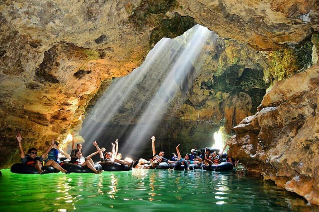 A group floats on rubber tubes through Pindul Cave's underground river, lit by a sunbeam