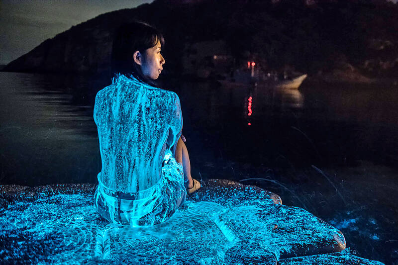Glowing blue bioluminescence in the water beside a person seated on the shore at night