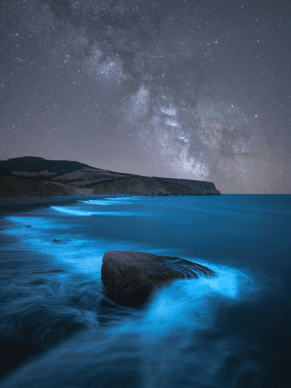 Bioluminescent blue water swirling past a rock beneath the Milky Way on a rocky coast
