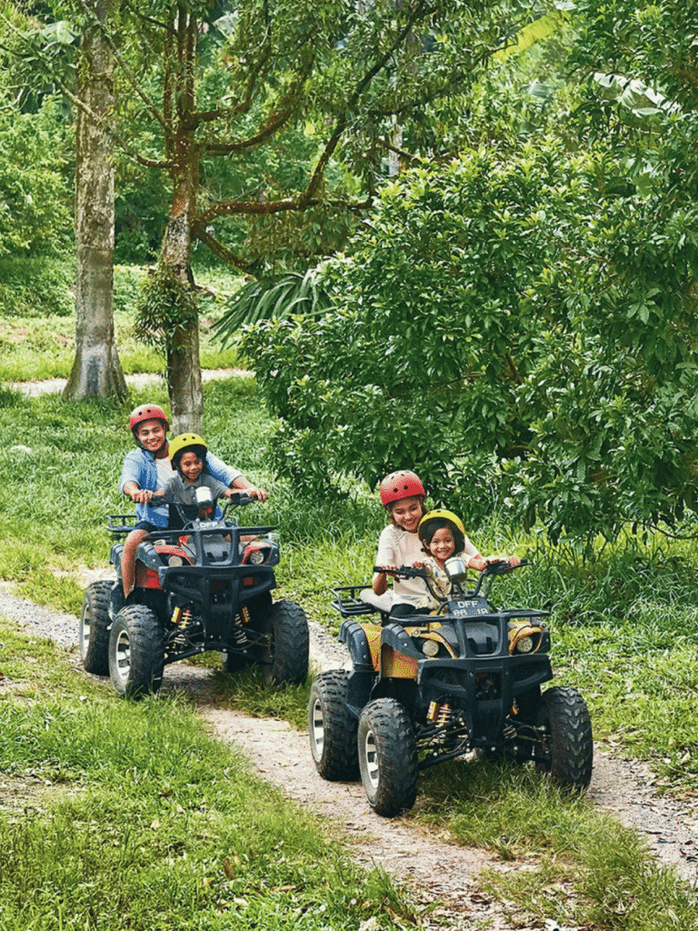 Two adults and two children in helmets riding ATVs along a grass track at Desaru Fruit Farm