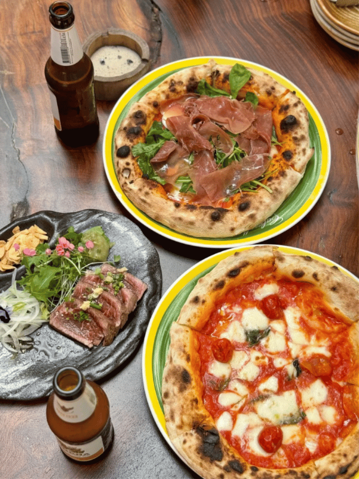 Two wood-fired Neapolitan pizzas, a sliced beef dish and beers on a wooden table