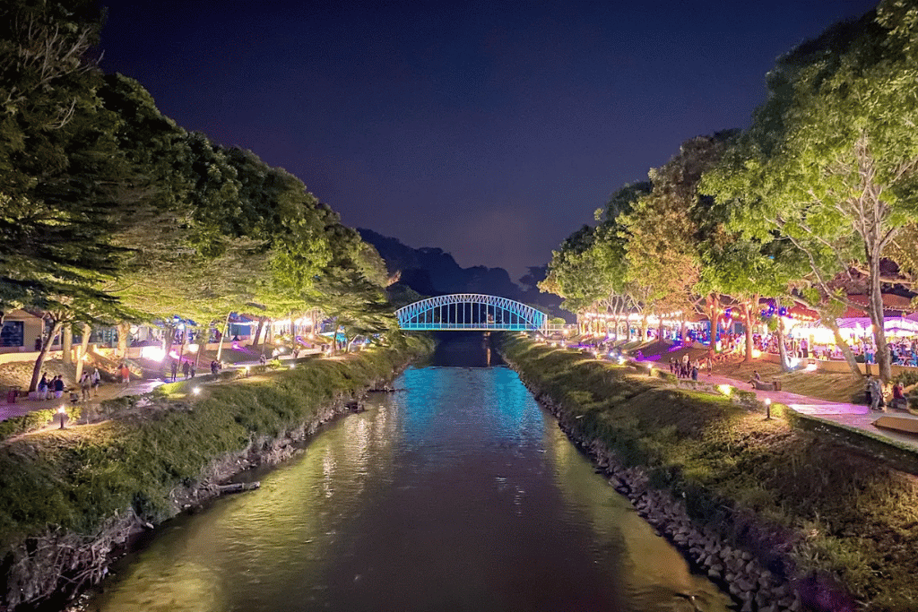 A lit arched bridge over the Kinta River at night with people along the illuminated banks