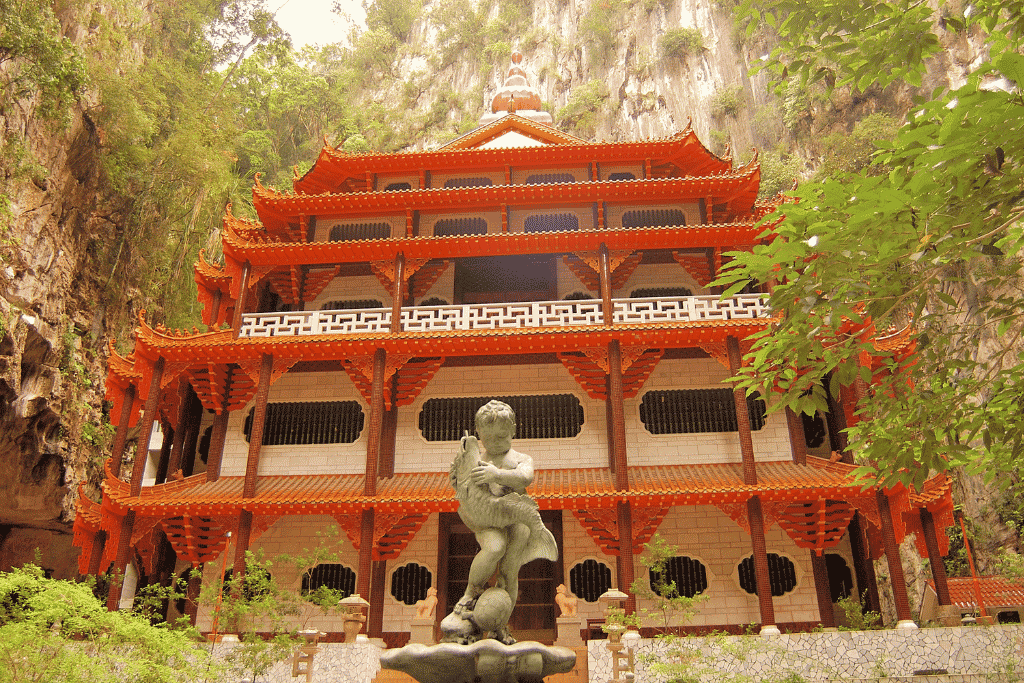 A tall red multi-tiered temple pagoda set against a jungle-clad limestone cliff in Ipoh
