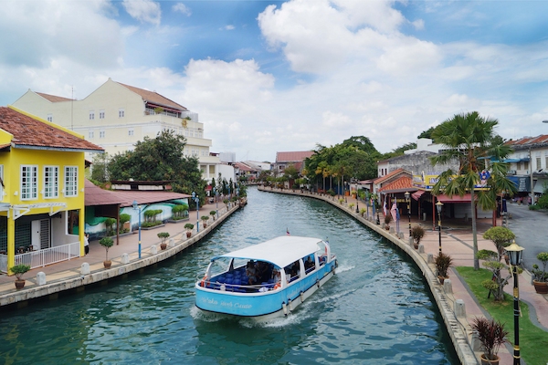 A blue river cruise boat heading down the Malacca River past colourful shophouses