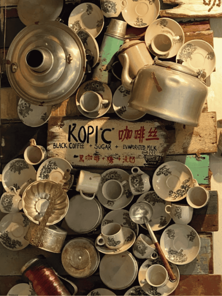 A wall of vintage teapots and cups around a retro KOPI-C coffee sign in an eclectic Malacca cafe