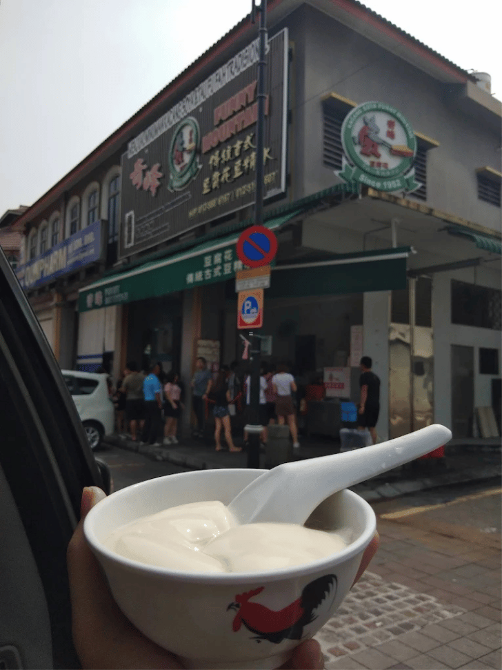 A bowl of silky tau fu fah held in front of the Funny Mountain soybean pudding shop in Ipoh