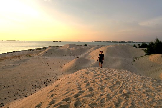 A person walking the pale sand dunes at Pantai Klebang as the sun sets over the strait