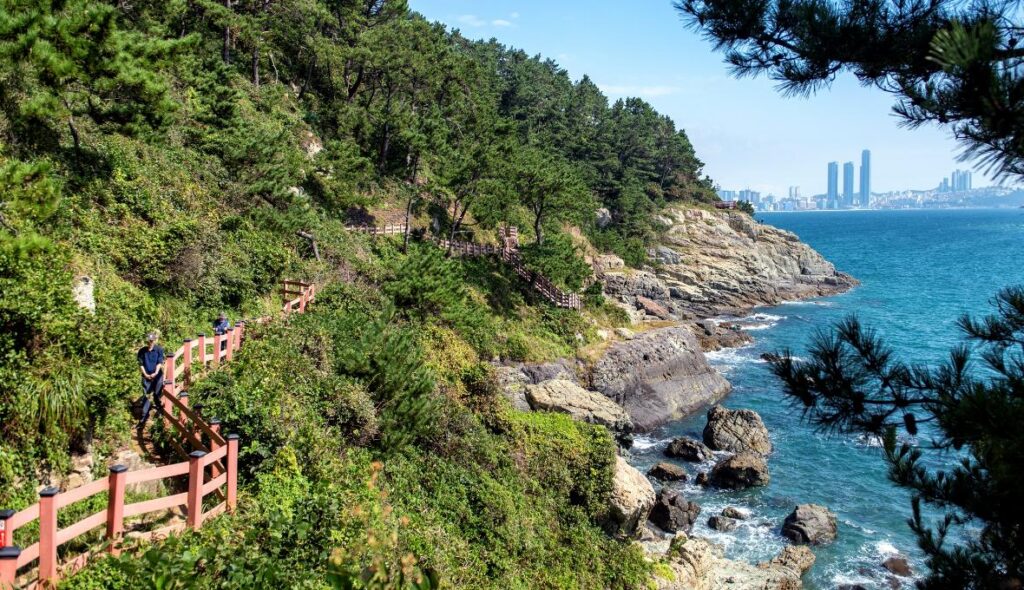 Wooden coastal trail with red railings, sea and distant city skyline at Igidae