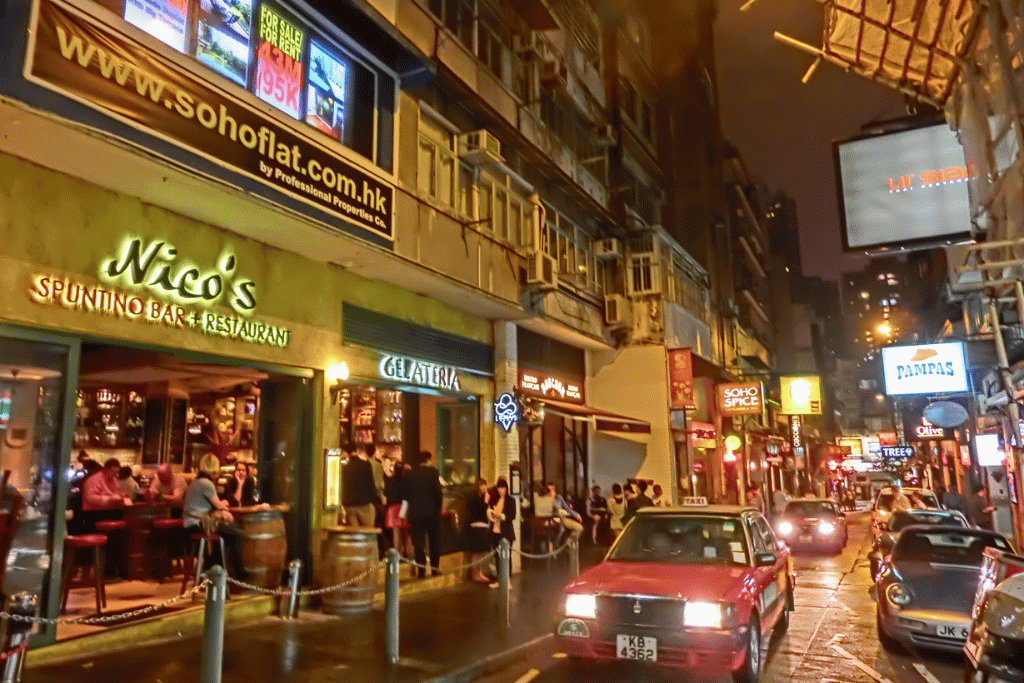 A red taxi on a neon-lit, rain-slicked street in Hong Kong's SoHo bar district at night