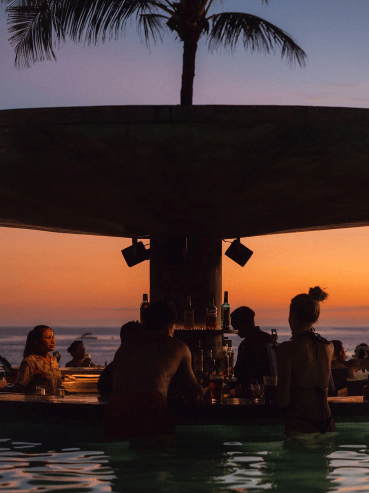 People at a beachfront pool bar under palms at sunset in Seminyak, Bali