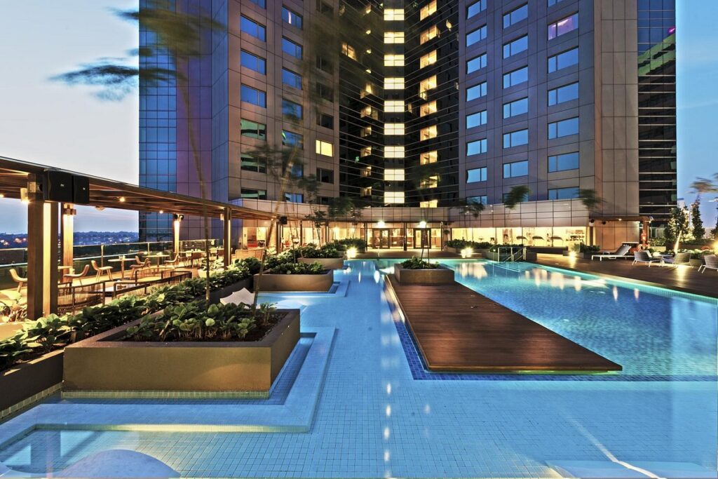 Landscaped rooftop hotel pool at dusk below lit glass towers in Johor Bahru