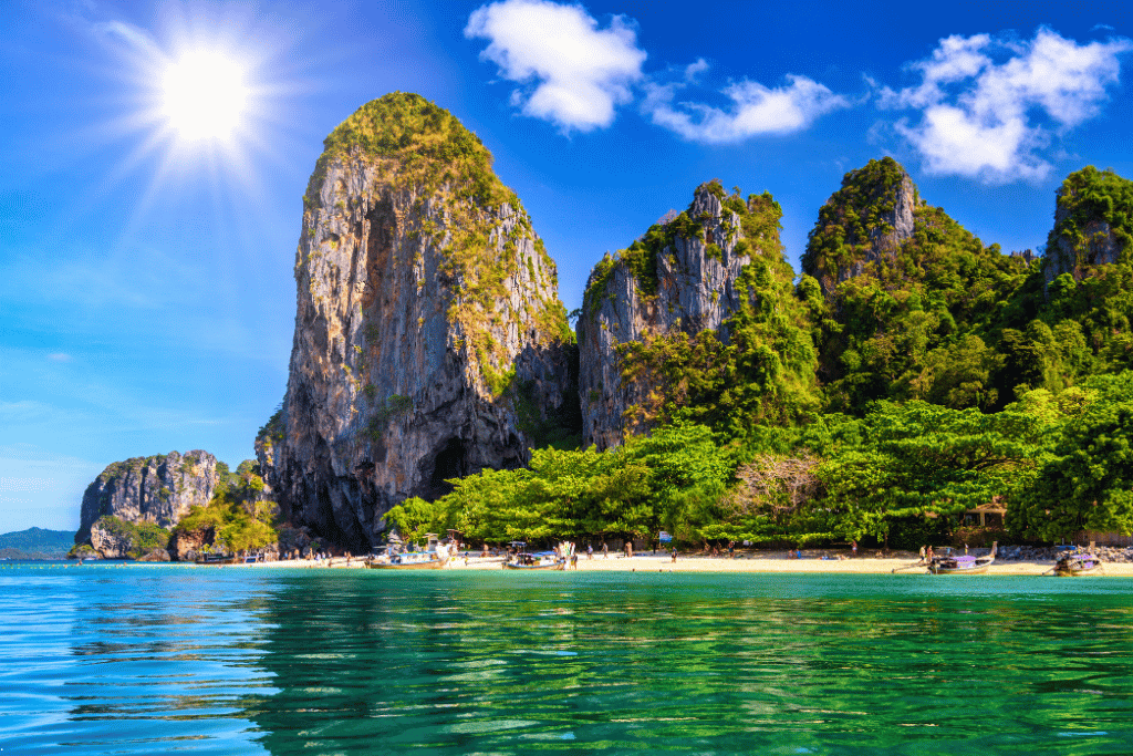 Sun over a towering limestone karst above a beach and turquoise water with longtail boats, Krabi