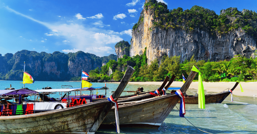 Longtail boats with Thai flags moored on a sandy beach beneath a limestone cliff, Krabi