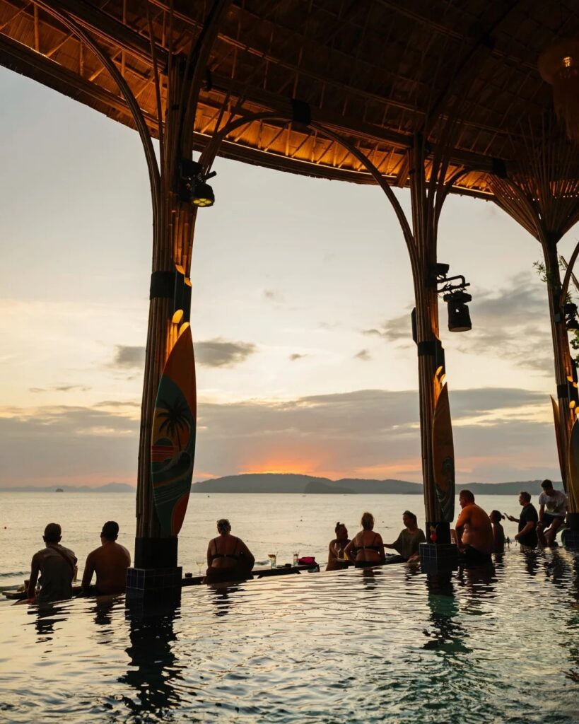Silhouetted beachfront bar with people in the water watching sunset over the sea, Ao Nang