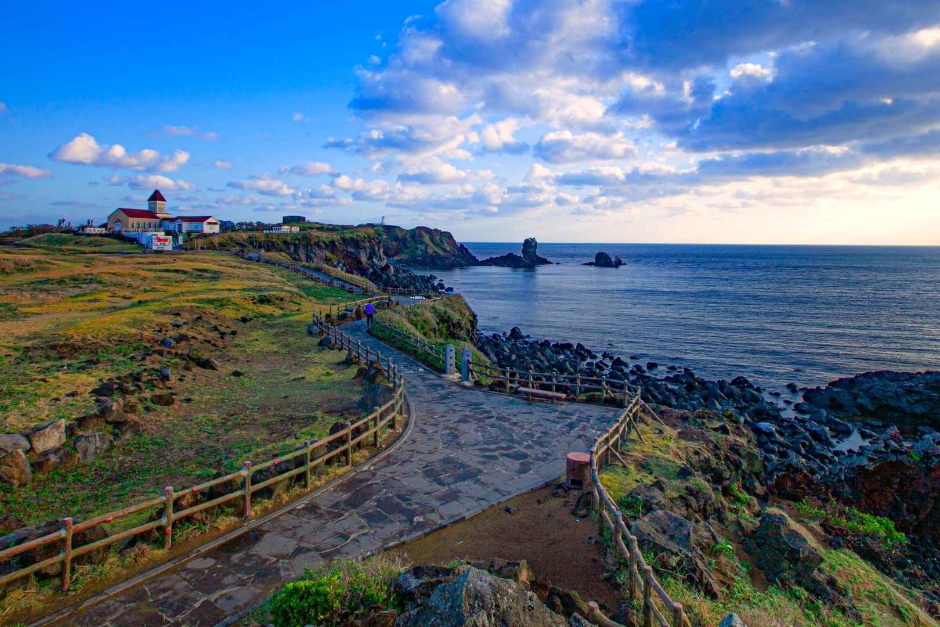 Coastal path along Seopjikoji cape past a red-roofed white building, with a sea stack offshore