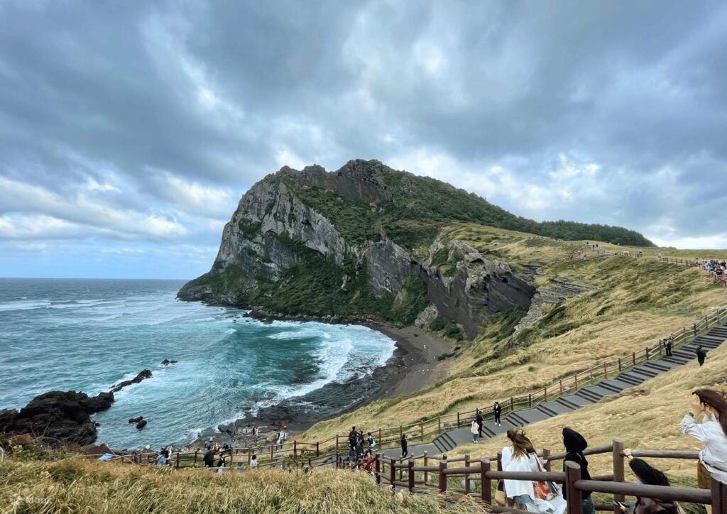 Visitors on a clifftop trail overlooking a green volcanic headland and sandy cove, east Jeju