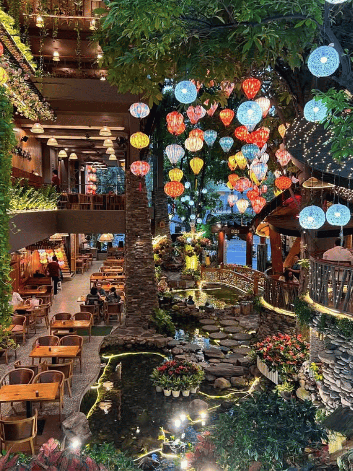 A lush, plant-filled garden cafe interior strung with colourful hanging lanterns
