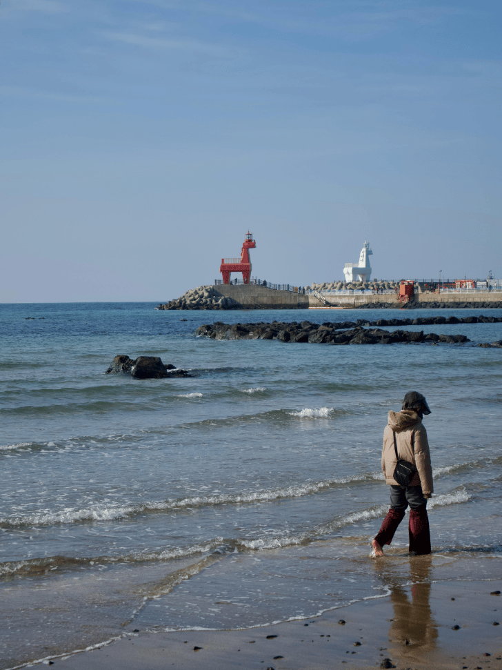 Person walking along a Jeju beach with red and white harbour lighthouses beyond