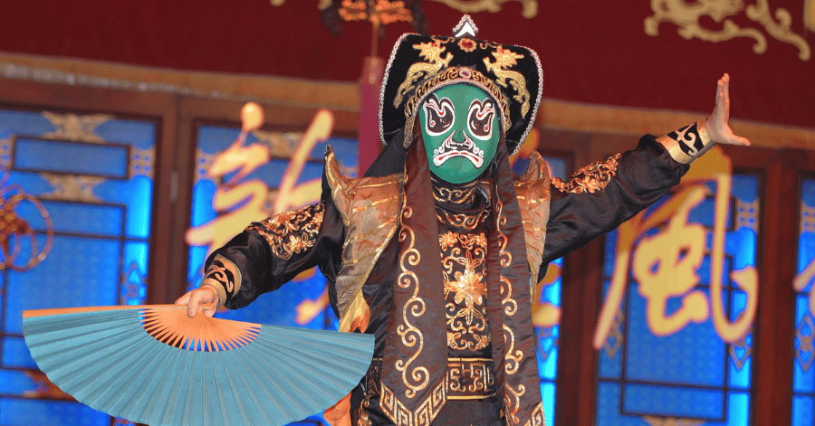 A Sichuan opera performer in a green face-changing mask and ornate costume, holding a fan