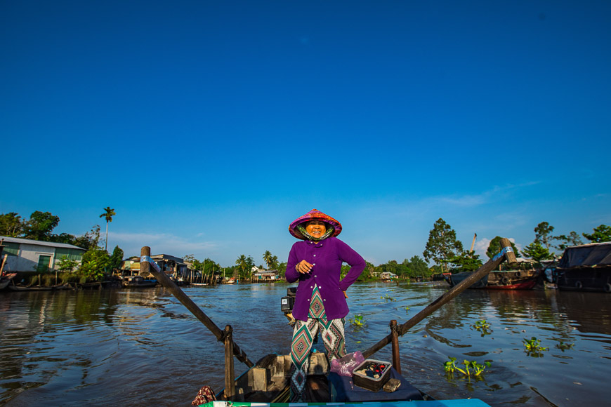 A woman in a conical hat rowing a wooden boat on a Mekong Delta river under blue sky