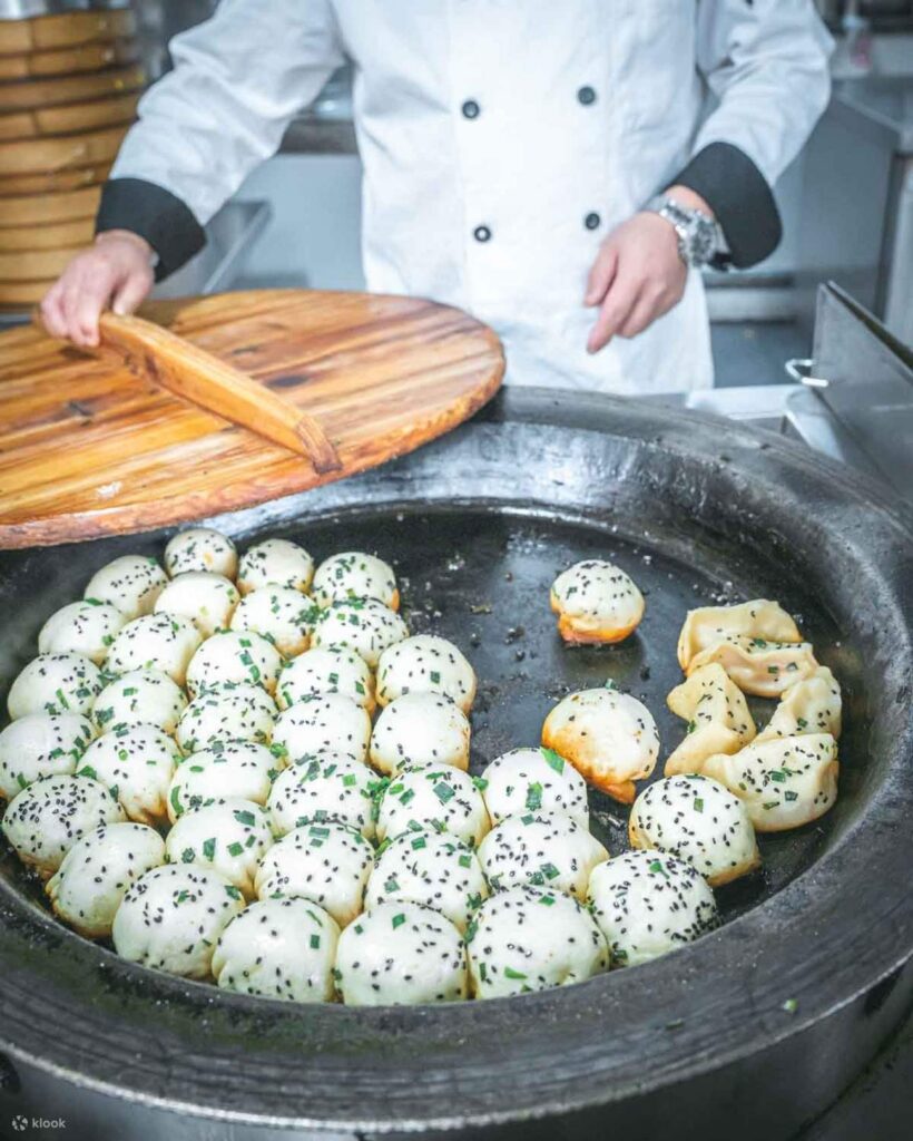 A cook lifting a wooden lid off a pan of sesame-topped pan-fried buns at a Shanghai stall