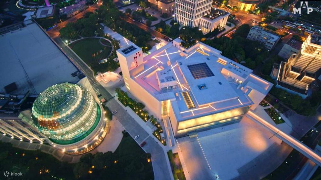 Aerial night view of the angular Museum of Art Pudong beside a lit glass dome in Shanghai