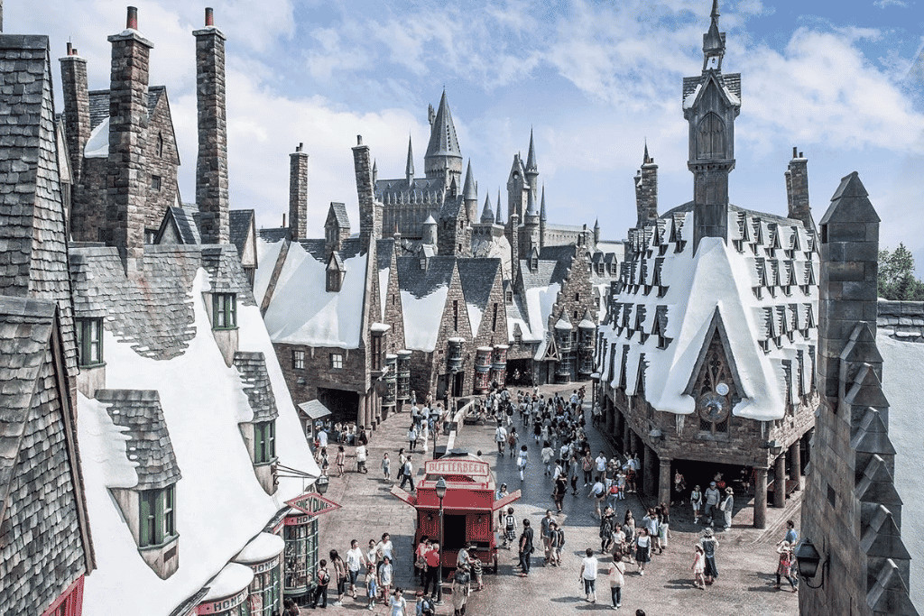 Snow-topped Hogsmeade rooftops with Hogwarts Castle behind and crowds by the Butterbeer cart