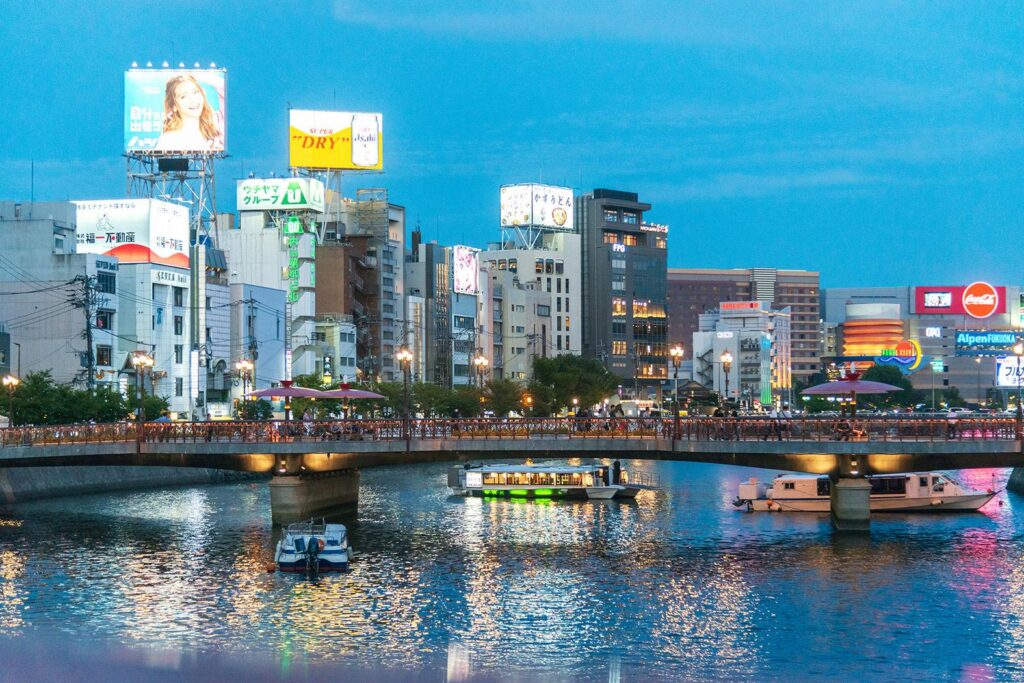 Nakasu's neon-lit riverside at dusk, with billboards, a bridge and boats on the water in Fukuoka