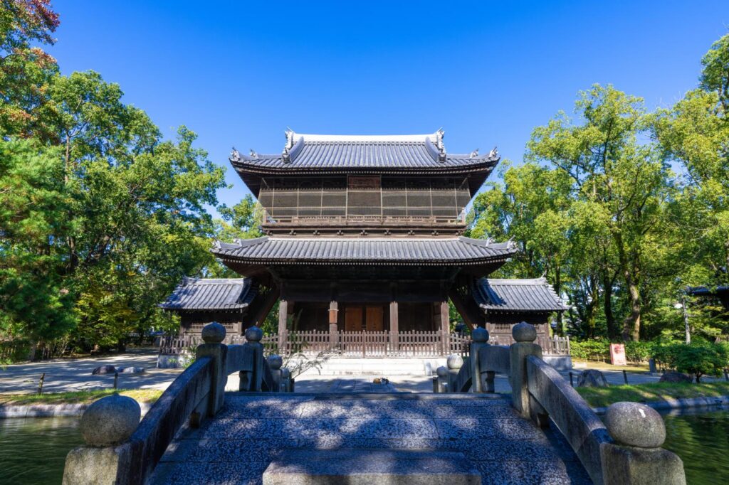 The two-storey wooden Sanmon gate of Shofukuji temple beyond a stone bridge, framed by trees