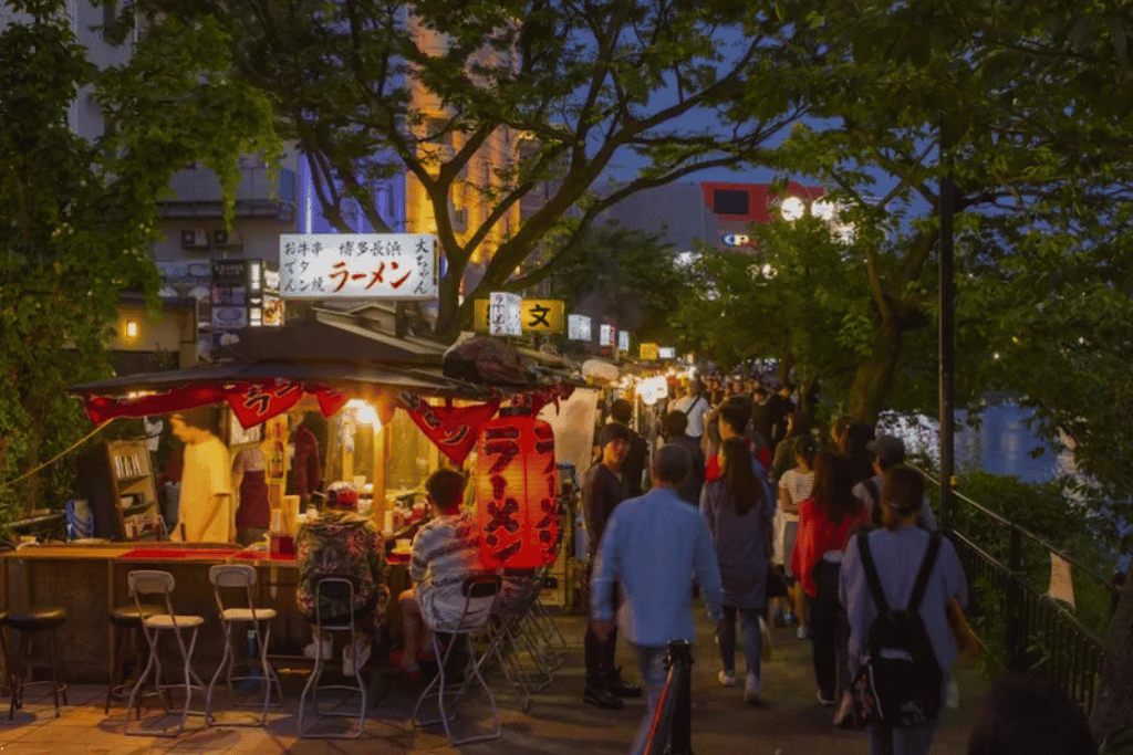 A lantern-lit yatai ramen stall at night with diners and a busy riverside crowd in Fukuoka