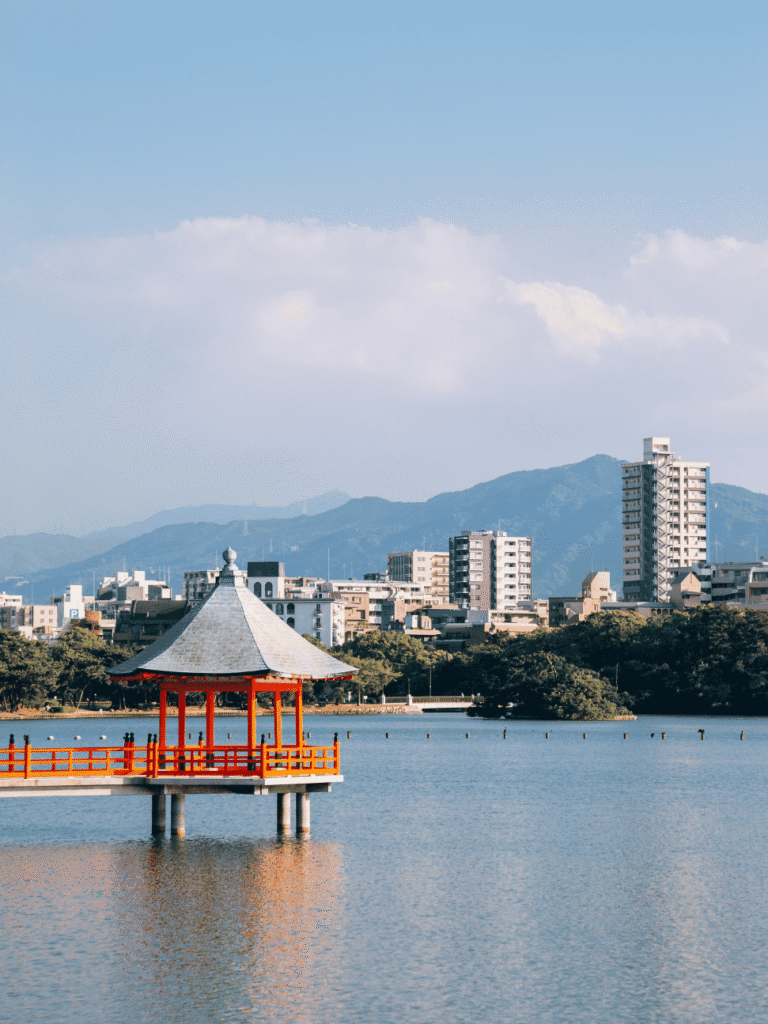 A red lakeside pavilion on Ohori Park's pond with the Fukuoka skyline and mountains behind