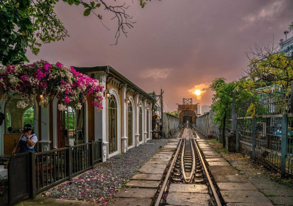Bougainvillea-covered cafe beside railway tracks leading to Long Bien Bridge at sunset